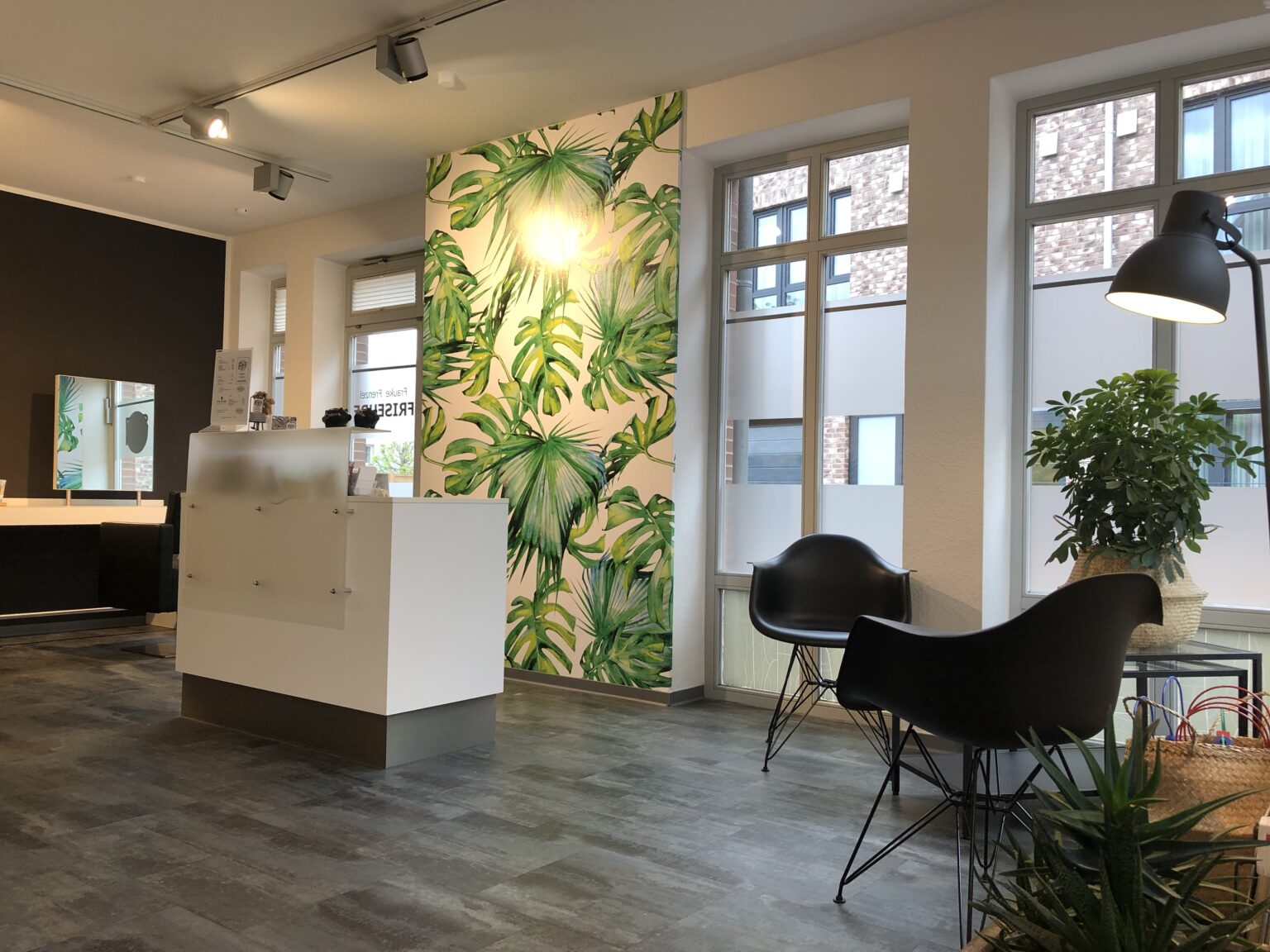 Heller, modern eingerichteter Friseurladen mit Empfangstresen, großen Fenstern und grauem Boden. Eine Wand ist mit einer grünen Blättertapete gestaltet, davor stehen schwarze Sitzstühle, Pflanzen und eine Stehlampe, die dem Raum eine ruhige, einladende Atmosphäre verleihen.